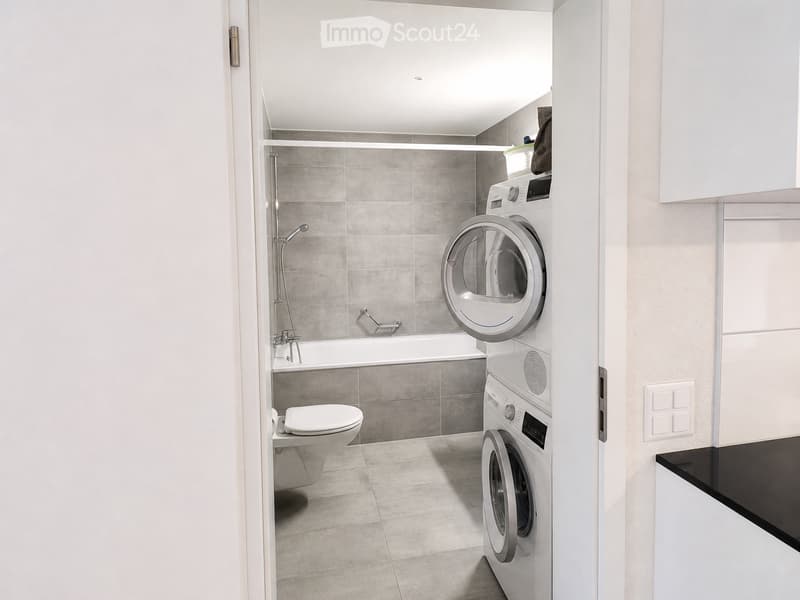 Modern bathroom with a toilet, bathtub, shower, and stacked washing machine and dryer unit.