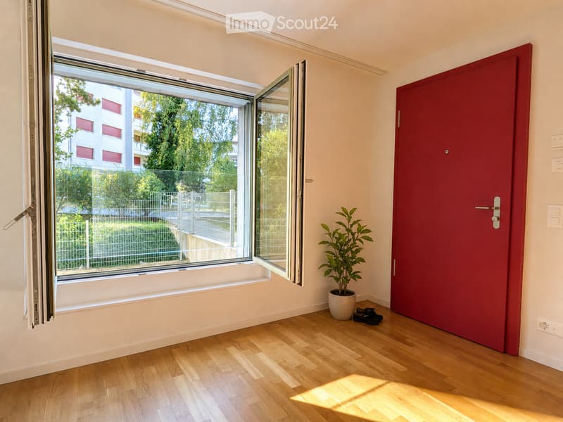 Empty room with wooden flooring, a large window with a view of greenery, a red door, a small potted plant, and a pair of shoes near the door.
