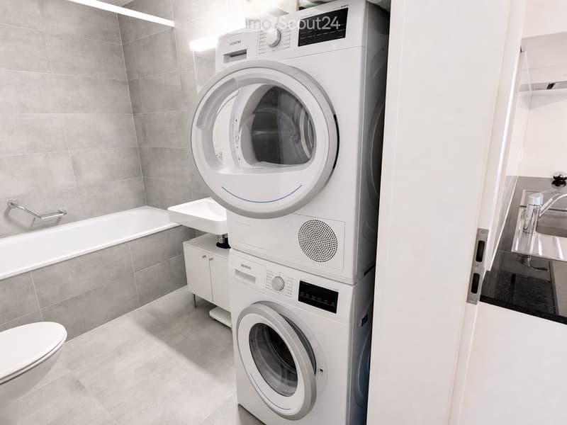 Modern bathroom with a white tiled wall, a bathtub, a toilet, a sink with a modern faucet, and a stacked Siemens washing machine and dryer unit.