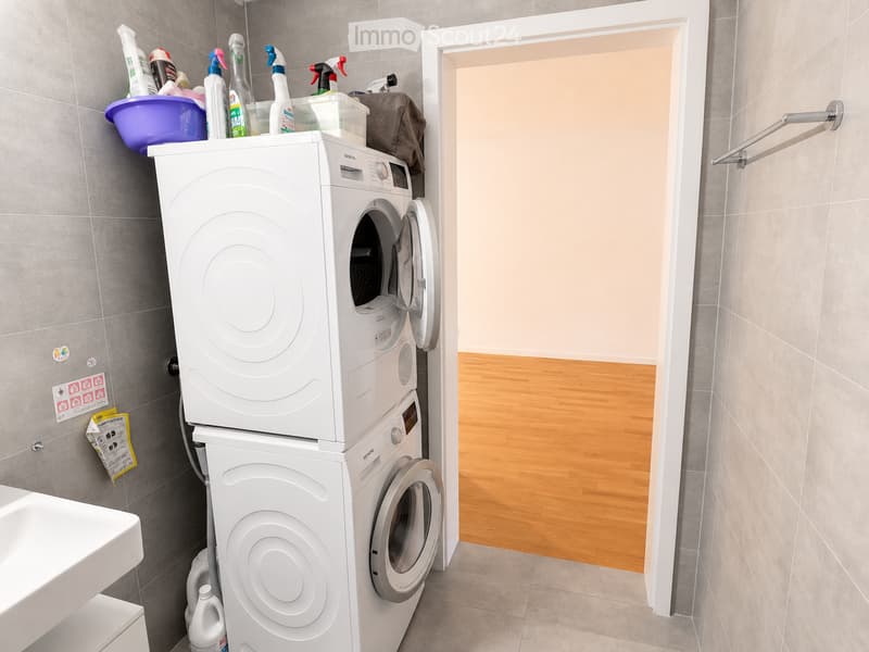 Modern bathroom with a white washing machine, sink, towel rack, and cleaning supplies on top of the washing machine.