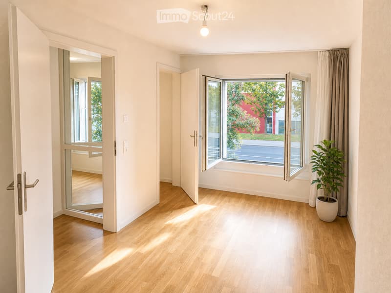 Empty room with light wooden flooring, white walls, large windows with open blinds, and a potted plant near the window.