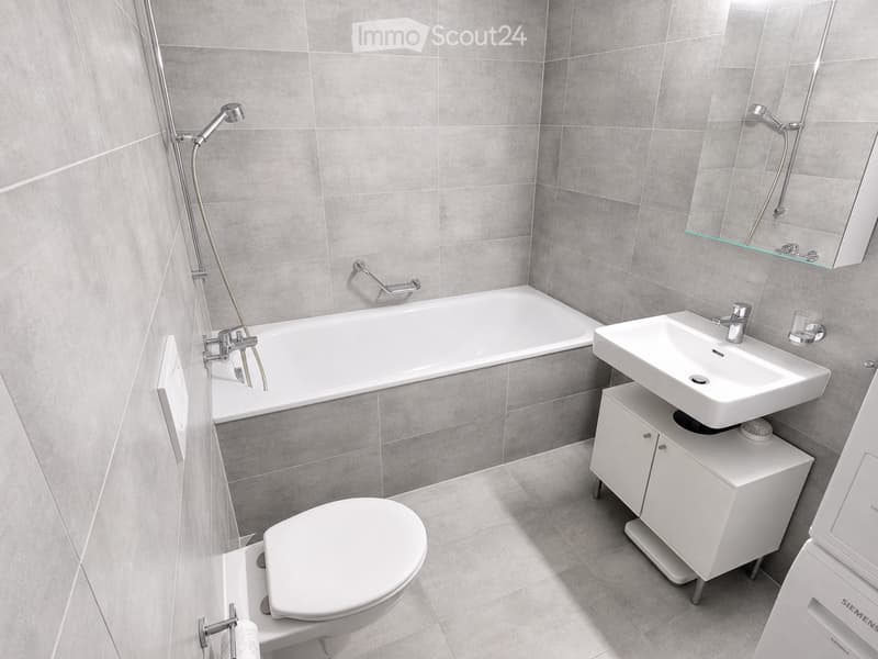 Modern bathroom with grey tiled walls and floor, featuring a bathtub, toilet, sink with cabinet, mirror, and showerhead.