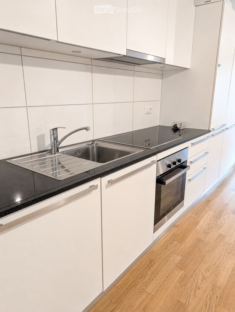 Modern kitchen with white cabinets, stainless steel sink, faucet, and oven. Black countertop and wooden flooring.