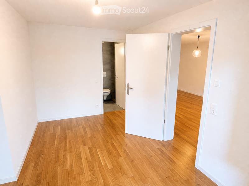 Empty hallway with wooden flooring, white walls, and two open doors leading to other rooms. One door leads to a small bathroom with a toilet, and the other leads to another room with wooden flooring and a hanging light fixture.