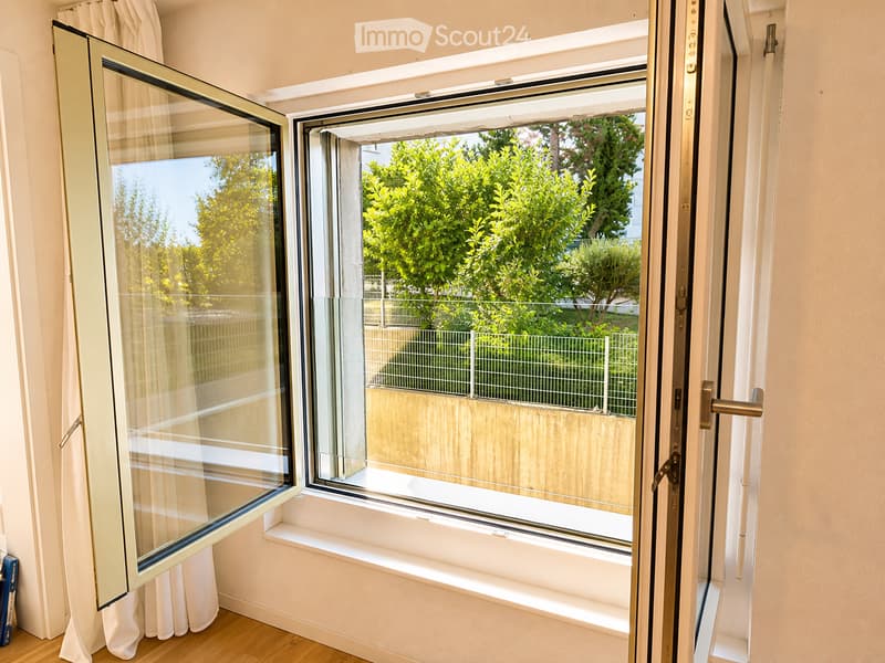 View of an open window and balcony door with a view of a garden area with greenery and a wooden fence.