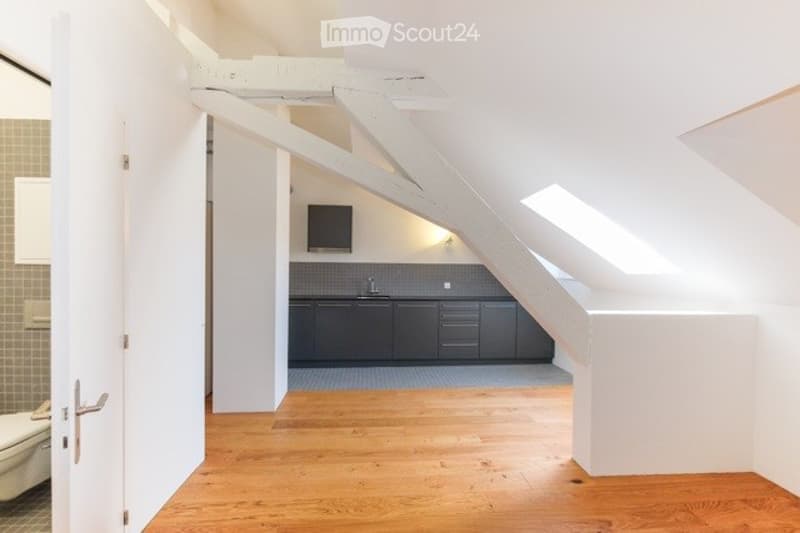 Loft-style kitchen area with grey cabinets, a sink, and a small screen mounted on the wall. The space features wooden flooring and exposed white beams.
