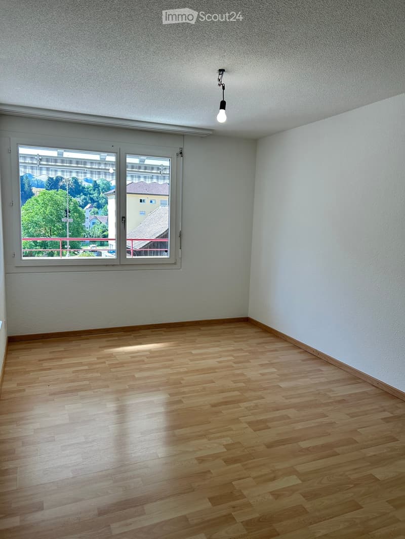 Empty room with wooden flooring, white walls, and large windows offering a view of the outside. A single pendant light hangs from the ceiling.