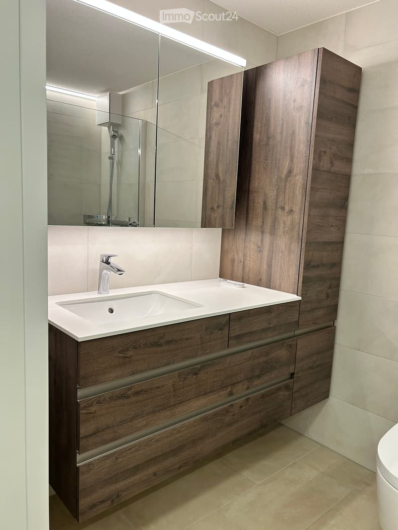 Modern bathroom with a wooden vanity, white countertop, sink, and mirror. Features a sleek faucet and built-in storage cabinets.