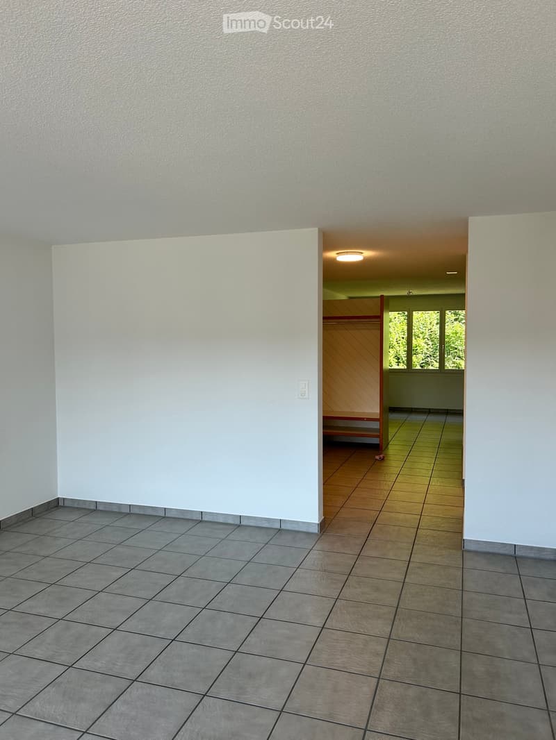 Empty room with tiled floor and white walls, featuring an open doorway leading to another room with wooden flooring and a window showing greenery outside.
