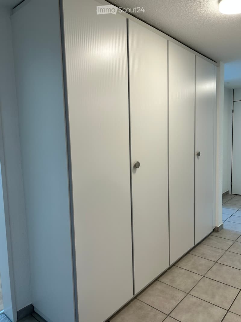 A narrow hallway with white paneled walls and tiled flooring, featuring closed white cabinet doors on one side.