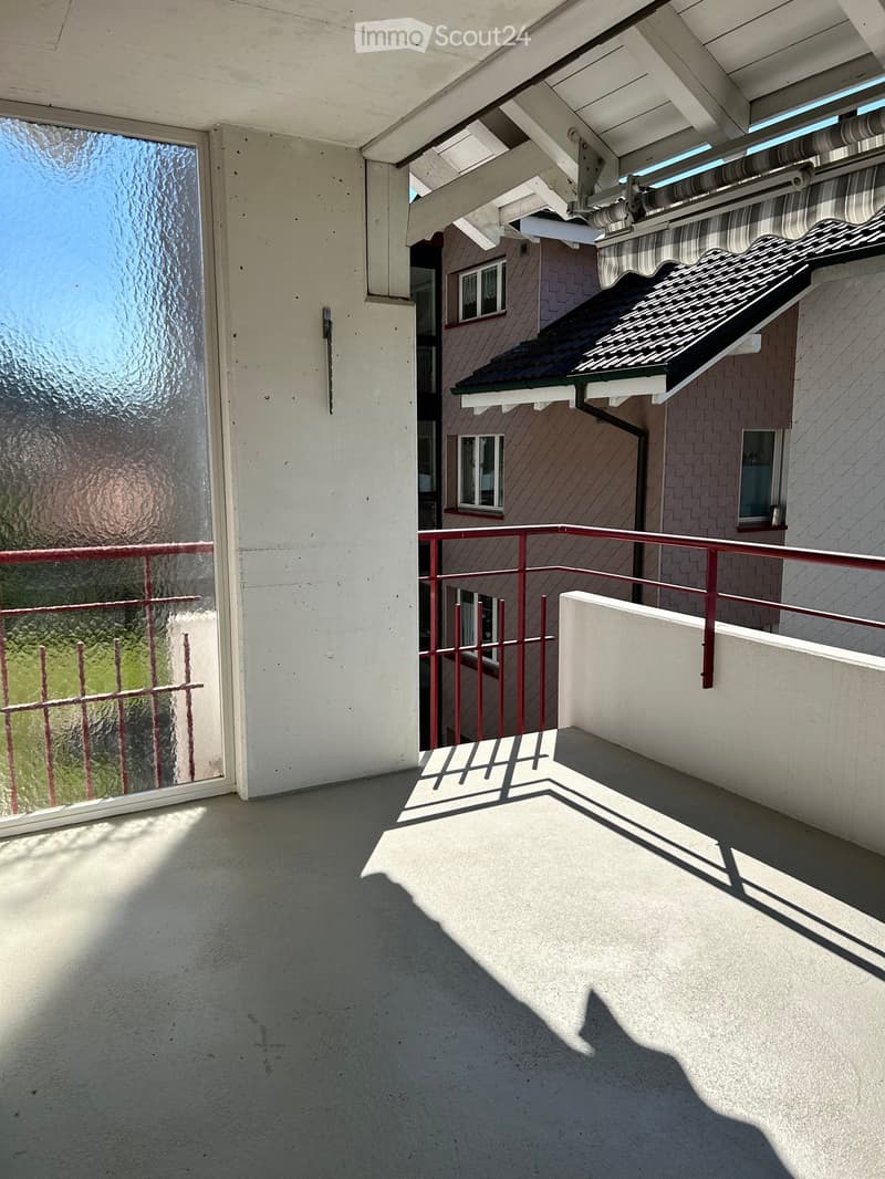 Balcony with red metal railing, glass door, and view of greenery. Retractable awning visible on adjacent building.