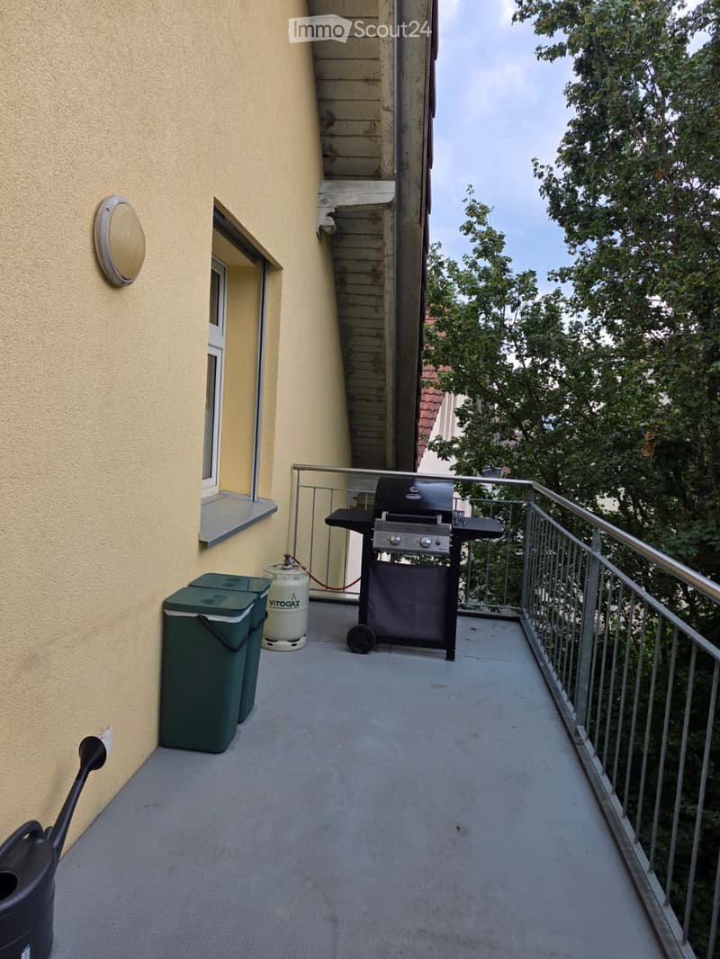 Balcony with a grill, two green trash bins, a gas cylinder, and a metal railing. The balcony is adjacent to a building with a staircase leading upwards.