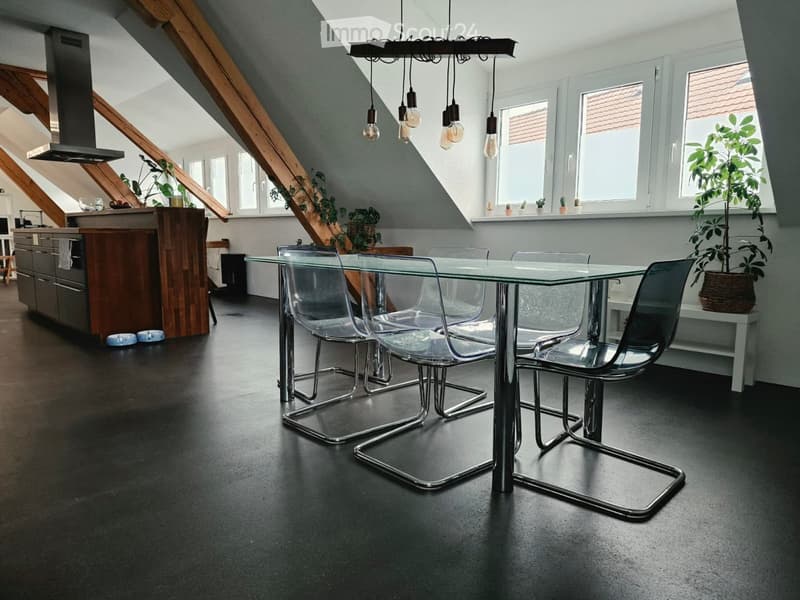 Modern open-plan kitchen and dining area with dark flooring, glass dining table, modern chairs, kitchen island, pendant lights, and plants.