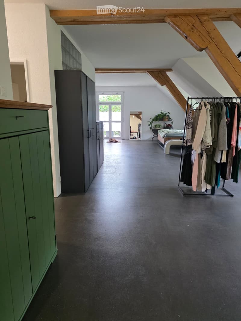 Spacious, open room with exposed wooden beams, dark grey flooring, and large windows. Features a wardrobe, clothing rack, bed, mirror, small table, and chair.