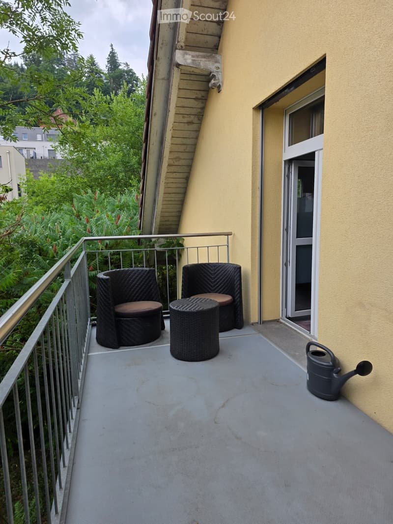 Balcony with two wicker chairs, a small table, and a watering can, overlooking a green area with trees.