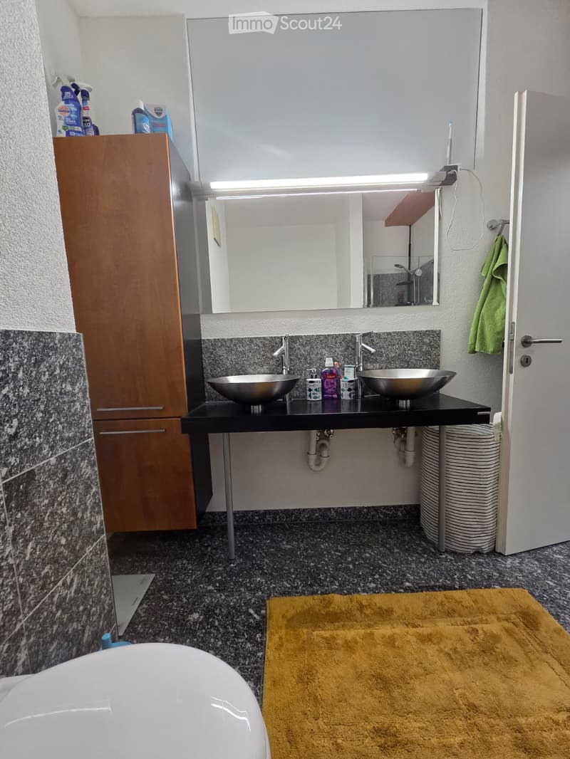 Modern bathroom with double sinks, granite countertops, a large mirror, a wooden cabinet, and a yellow bath rug.