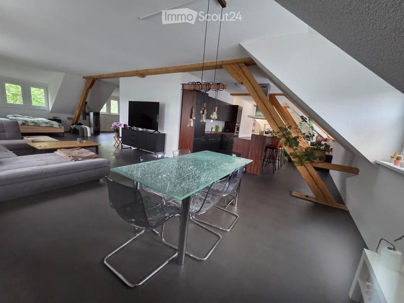Open-plan living and dining area with exposed wooden beams, grey sofa, coffee table, glass dining table with chairs, kitchen island, and a bedroom area with a bed and nightstand.
