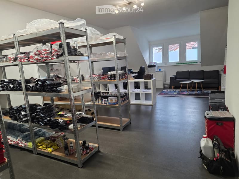 A well-lit room with metal shelving units filled with packaged items, a small seating area with a sofa and two stools, a table with a laptop, and a printer on a cabinet.