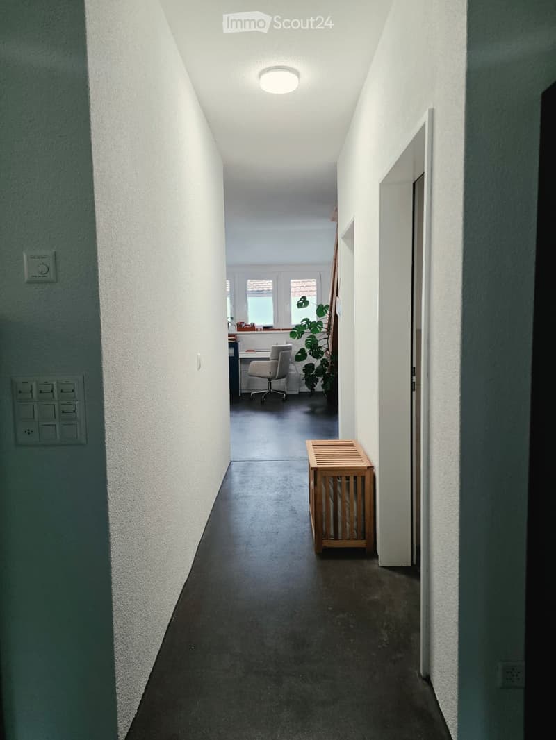 A narrow hallway with white walls, a dark floor, and a ceiling light. A small wooden bench is placed against the wall, and a glimpse of an adjacent room shows a desk, chair, and plant.