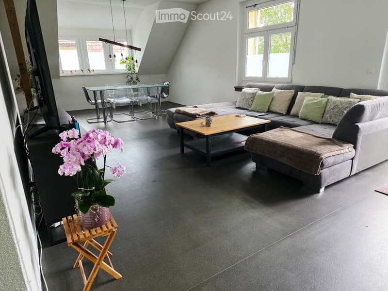 Modern living room with a grey sectional sofa, wooden coffee table, glass dining table, and a small wooden stool holding a flower pot. The room features large windows and a slanted ceiling.