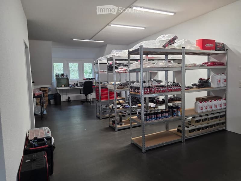 A well-lit storage or workshop room with metal shelving units filled with various items, including boxes, cans, and storage bins. A desk with a laptop and chair is visible in the background, along with additional storage items and a suitcase on the floor.