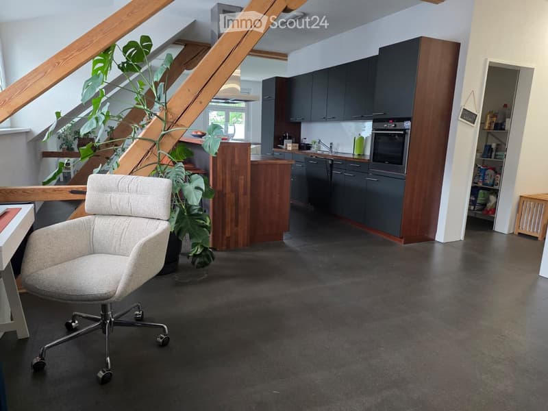 Modern, open-plan living and kitchen area with grey cabinets, a wooden island, a grey office chair, and a large leafy plant. The space features a dark grey concrete floor and a staircase leading upwards.