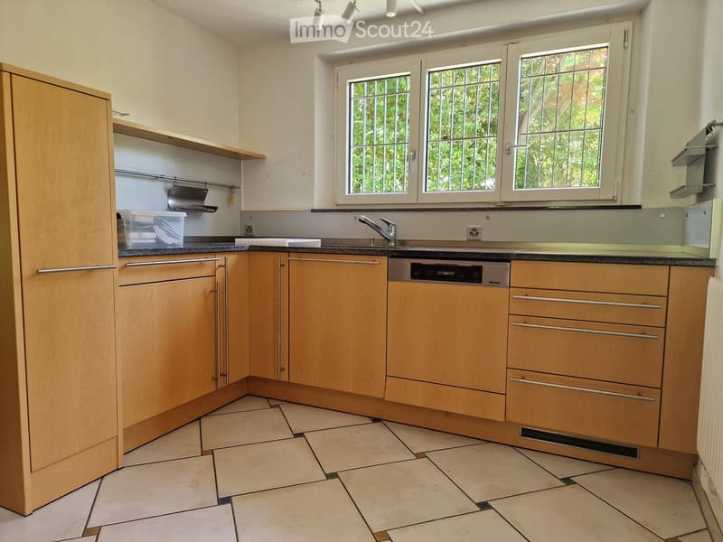 Modern kitchen with light wooden cabinets, tiled floor, sink, faucet, and dishwasher. Windows with white frames and metal bars provide natural light.