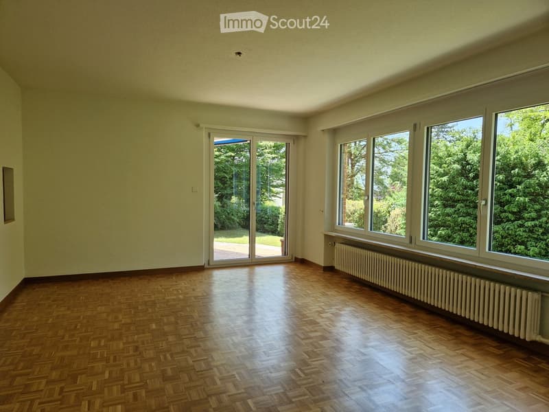 Empty room with wooden herringbone flooring, large windows, and a sliding glass door leading to a garden.