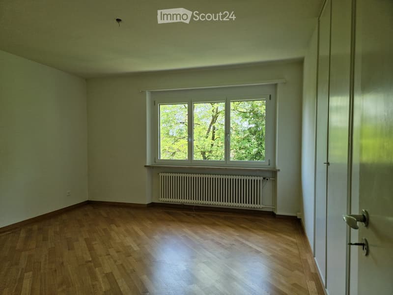 Empty room with wooden flooring, large window with white frame, radiator below window, and white walls.
