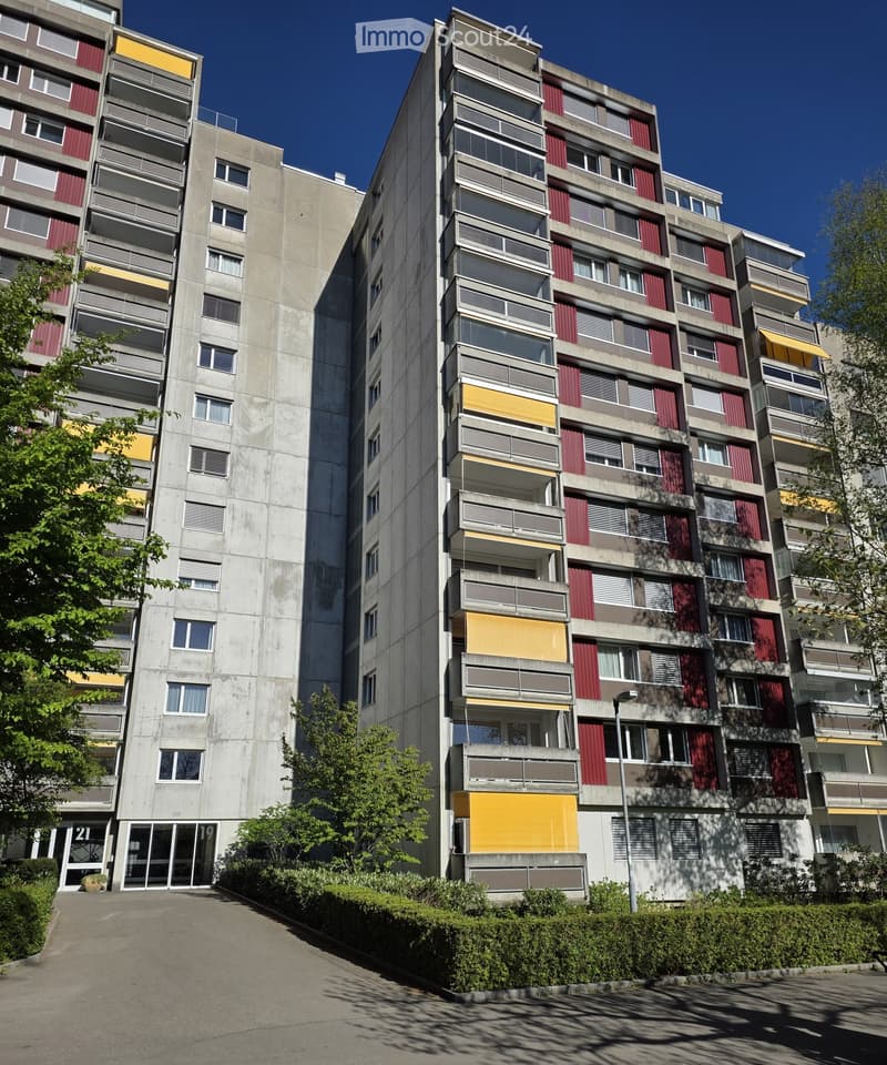 Multi-story residential building with concrete facade, red and yellow accents, multiple windows, and balconies.