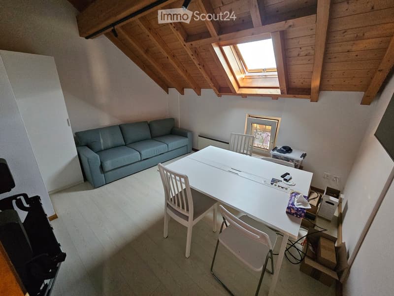 A furnished attic room with a grey sofa, a white dining table, and chairs, featuring exposed wooden beams and a skylight window.