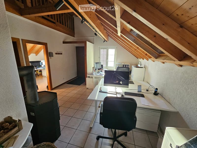 Office space with a wooden slanted ceiling, tiled floor, and large window. Includes a desk with a computer, chair, keyboard, and printer, along with a cabinet and additional workspace in the background.