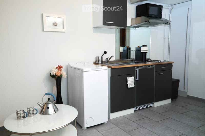 Compact kitchen with dark cabinets, stainless steel appliances, a small round table with teapot and cups, and a vase with flowers.