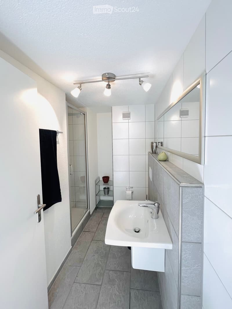 Modern bathroom with white tiles, a sink with a chrome faucet, a large mirror, a black towel, a glass shower enclosure, and a small metal shelf holding cups and a decorative vase.