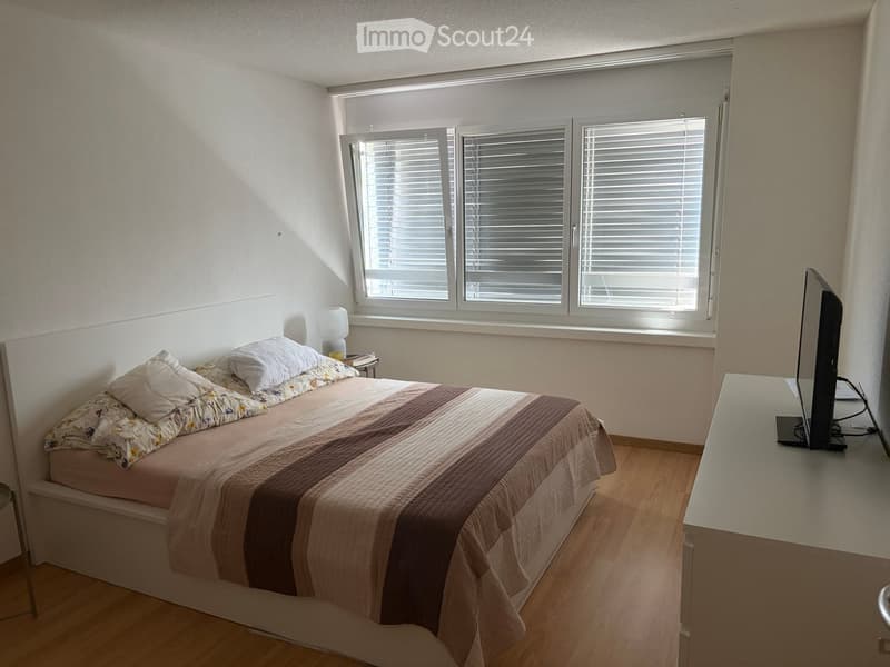 A well-lit bedroom with a bed, nightstand, lamp, and a screen on a table. The room features wooden flooring and large windows with blinds.