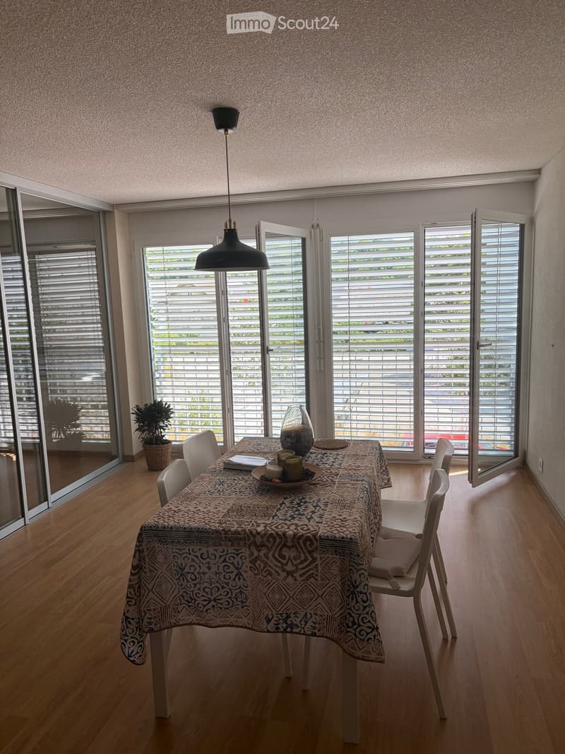 Bright dining room with wooden flooring, large windows with blinds, a dining table with chairs, a patterned tablecloth, a vase with candles, and potted plants.