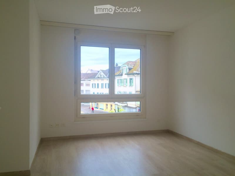 Empty room with wooden flooring and large windows offering a view of neighboring buildings.