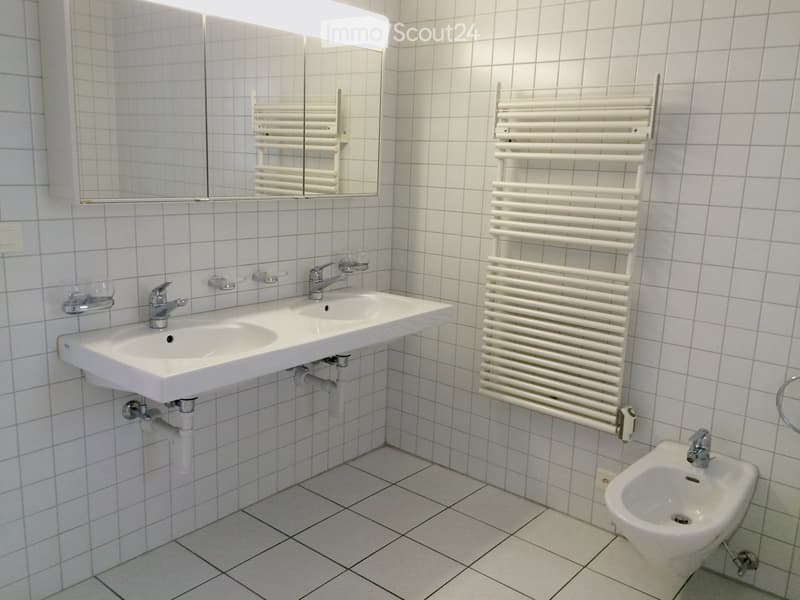 Modern bathroom with white tiled walls and floor, double sink, mirror, radiator, and bidet.