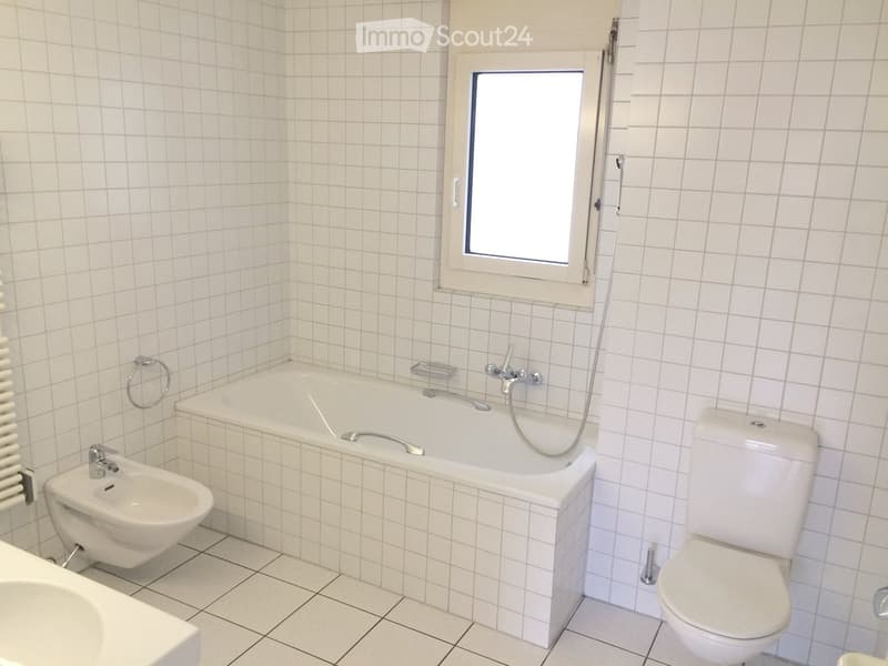Modern bathroom with white tiled walls and floor, featuring a bathtub, toilet, bidet, and a small window above the tub.