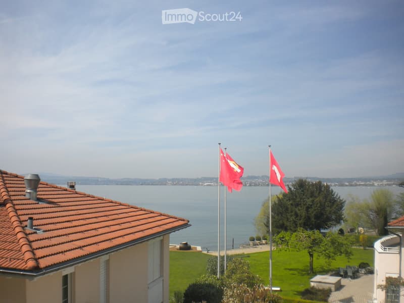 View of a house with a red-tiled roof, overlooking a body of water with flags on poles, surrounded by a well-maintained lawn and trees.