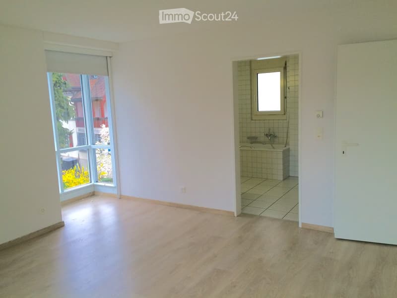 Empty room with wooden flooring, white walls, large window with blinds, and an adjacent small tiled bathroom with a bathtub and window.