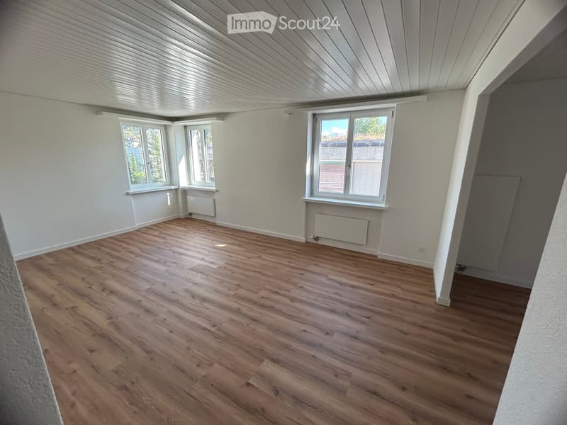Empty attic room with wooden flooring, white walls, and multiple windows offering natural light. The ceiling has a slanted design typical of attic spaces.