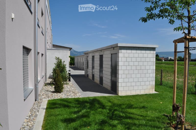 Two buildings, one gray and one white, gravel pathway, greenery, building with windows, building with doors, large grass field in the distance.