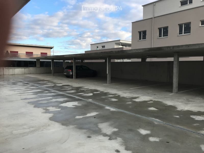 A vacant parking lot with concrete ground, two concrete supports, a parked car, and a building on the right side.