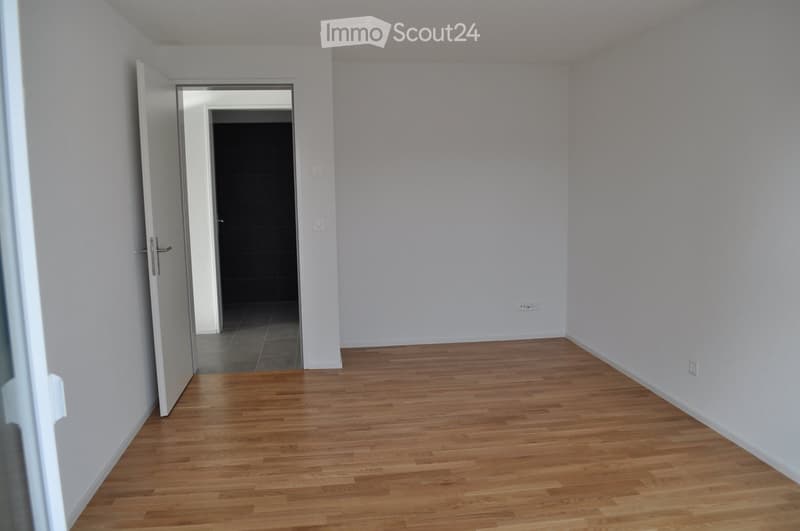 Empty room with white walls, wooden floor, open door, and interior door leading to a shower room. Outlets are on the walls.