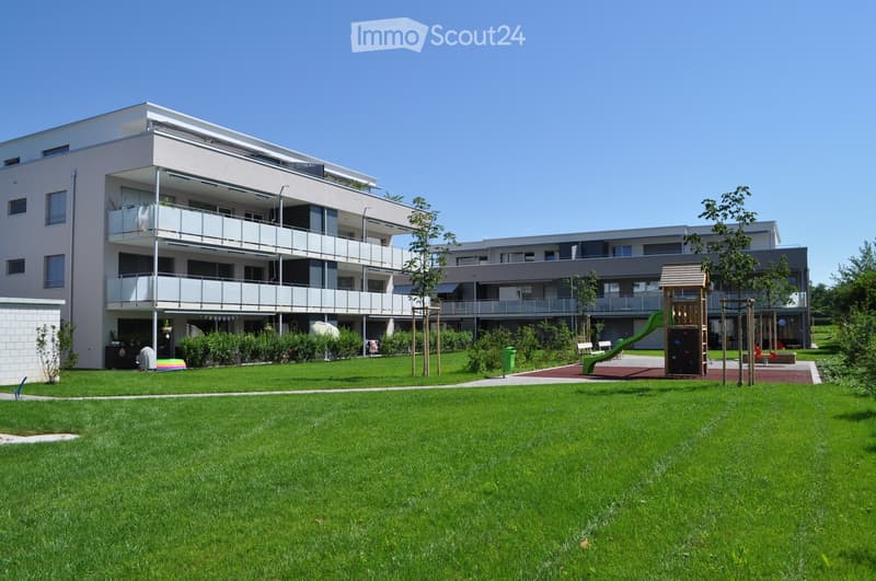 White apartment building with three floors, balconies, playground, grass lawn, plants, trees, and green slide.