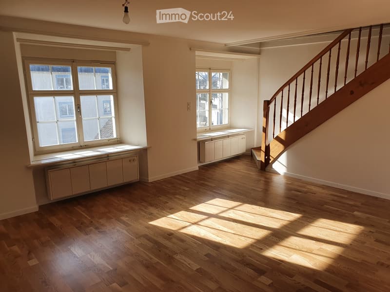 Empty room with wooden flooring, large windows, radiator, and staircase leading upwards.