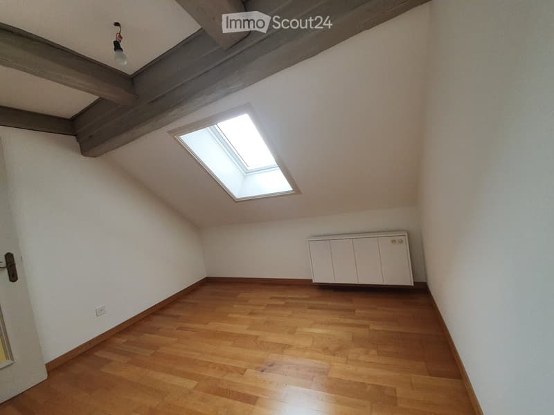 Empty attic room with wooden floor, white walls, exposed wooden beams, skylight window, and a small white cabinet.