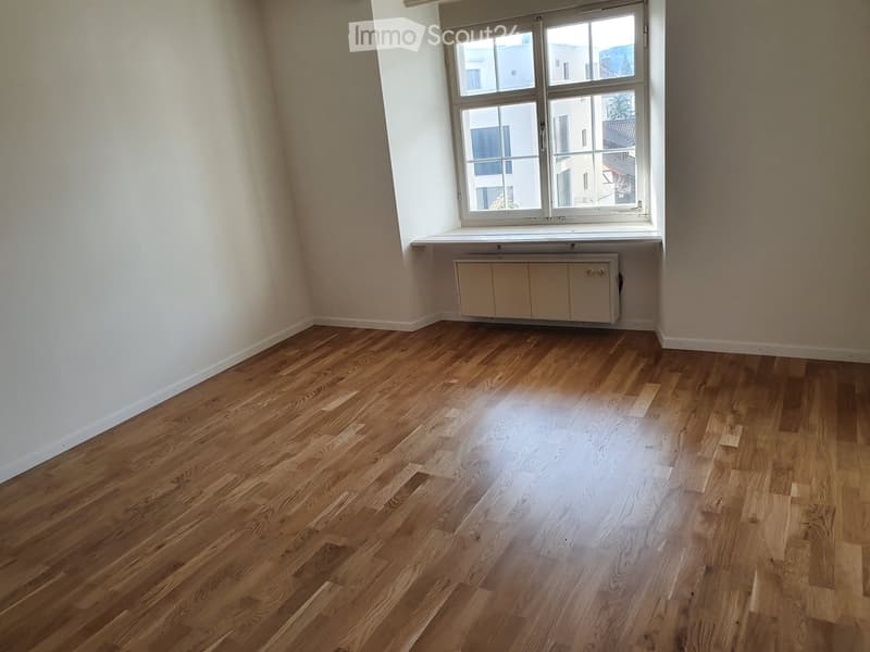 Empty room with wooden flooring, white walls, and a window with a radiator below it.