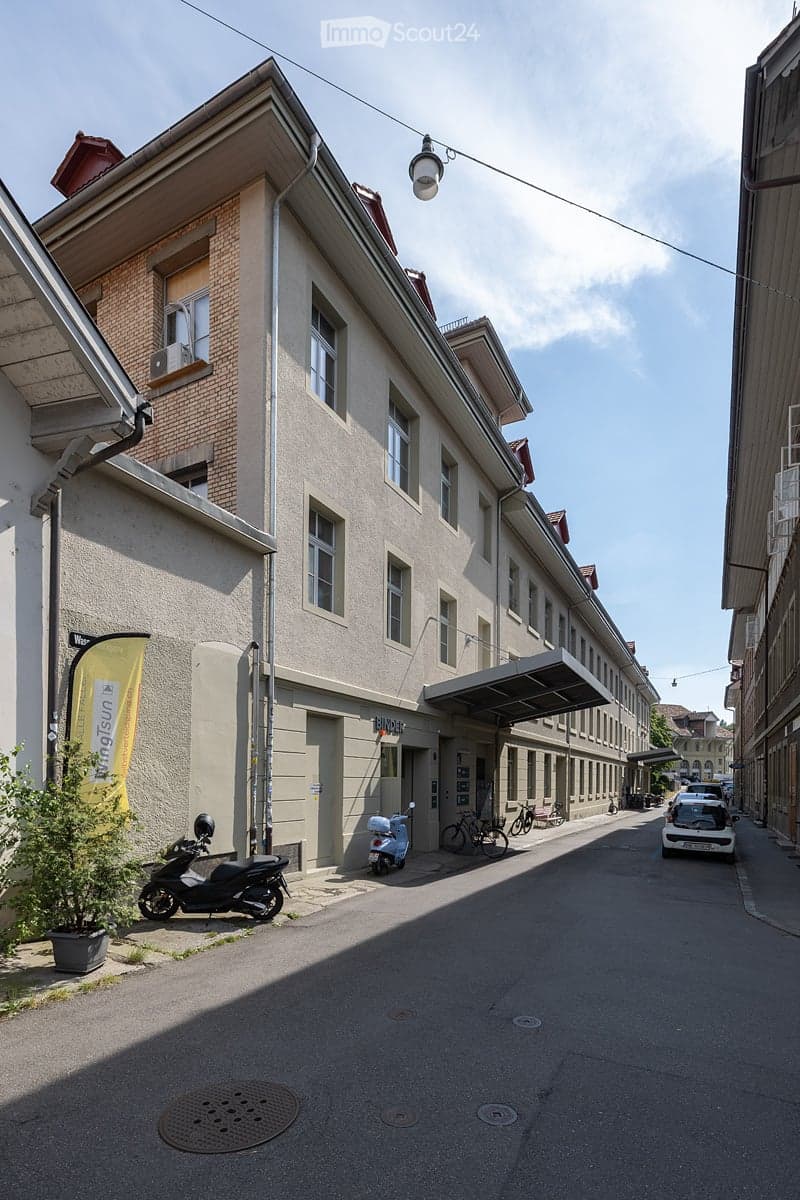 A multi-story building with a mix of brick and plaster facades, featuring several windows and a covered entrance. The street in front has parked vehicles, including a scooter and bicycles, and is lined with additional buildings.